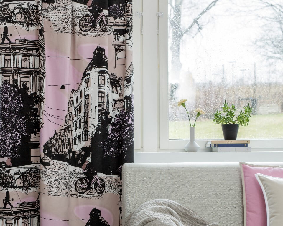 Wisząca przy oknie gotowa zasłona zaciemniająca BULEVARDI beżowa z obrazem ulicy Helsinek w aranżacji z sofą, poduszką i narzutą