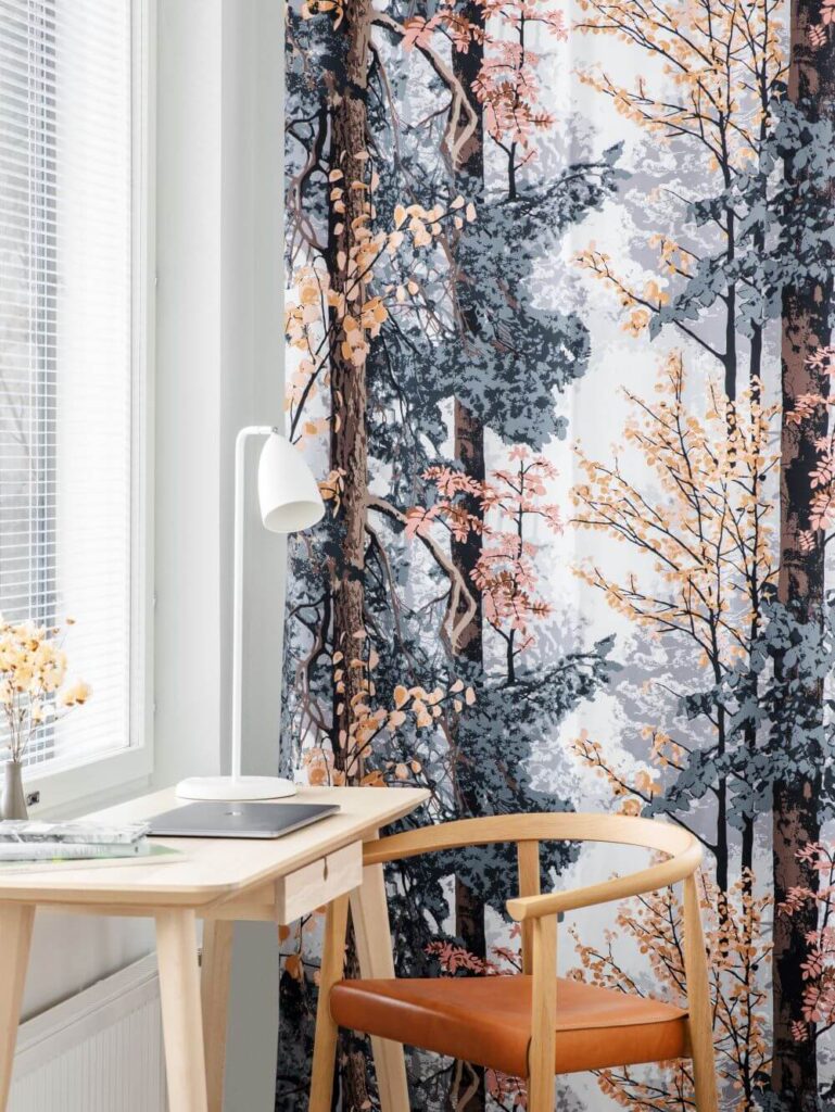 Kolorowe zasłony w liście na tle okna pięknie dekorują nowoczesny salon. Motyw liści, drzew i połączenia kolorów granatu, beżu, pomarańczowego na zasłonach dodają salonowi unikatowego skandynawskiego charakteru.