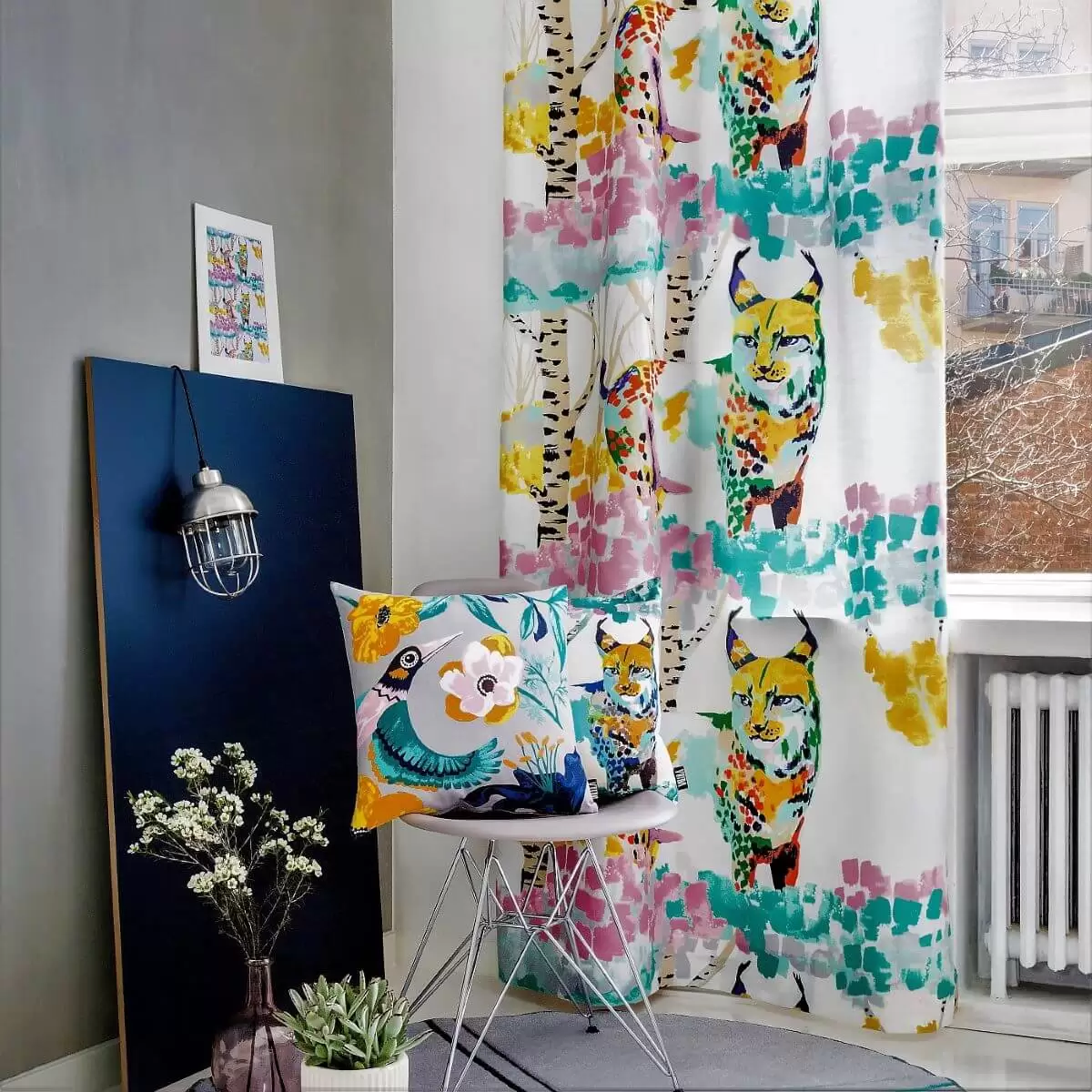Zasłona kolorowa LUMI ILVES w malowany nowoczesny leśny krajobraz z kolorowym rysiem w aranżacji w skandynawskim szarym salonie. Widok gotowej zasłony na taśmie zawieszonej przy białym oknie. Widoczny skandynawskie krzesło z kolorowymi poduszkami, fragment nowoczesnego dywanu, lampa oraz kwiaty