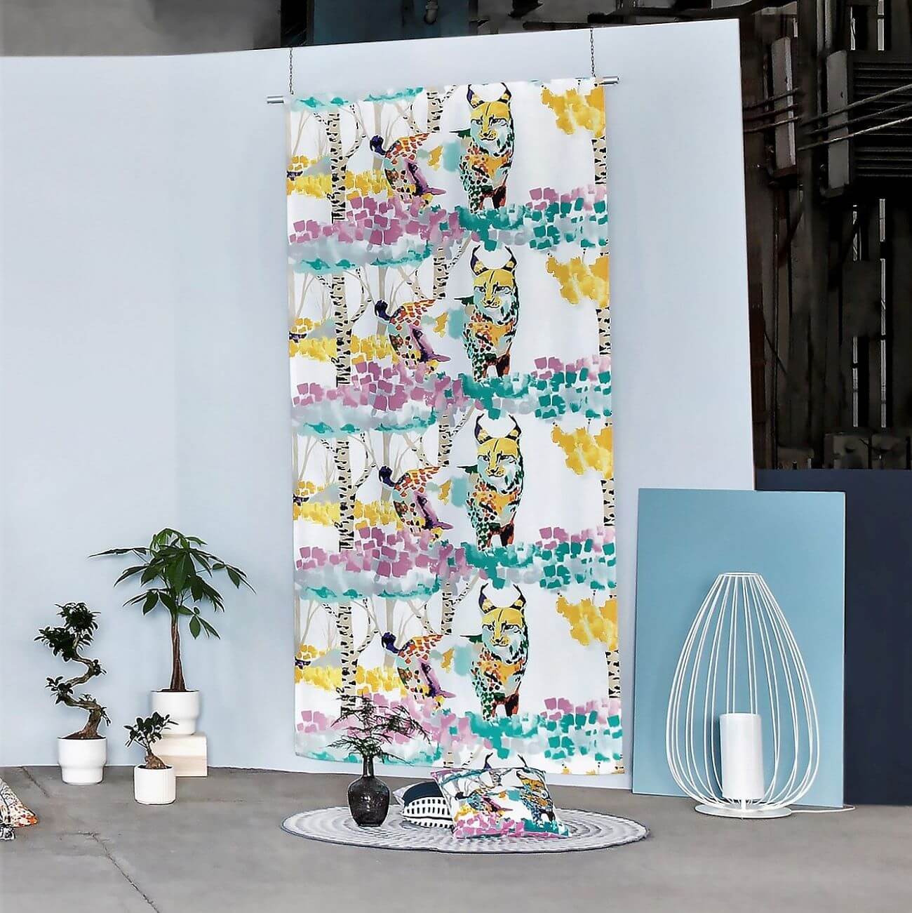 Zasłona kolorowa LUMI ILVES w malowany nowoczesny leśny krajobraz z kolorowym rysiem w aranżacji w studiu. Widok gotowej zasłony na taśmie dla dzieci przywieszonej na tle jasno niebieskiej ściany. Widoczny skandynawskie dywan, kolorowe poduszki, nowoczesna lampa oraz 4 kwiaty