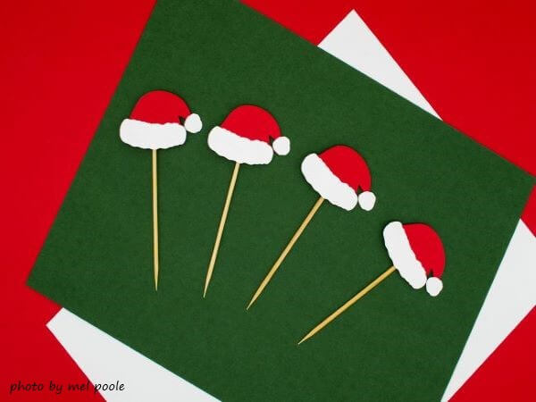 Własnoręcznie wykonana kartka z życzeniami świątecznymi. Na zielonym kartonie umieszczone są czerwone czapeczki świętego mikołaja.