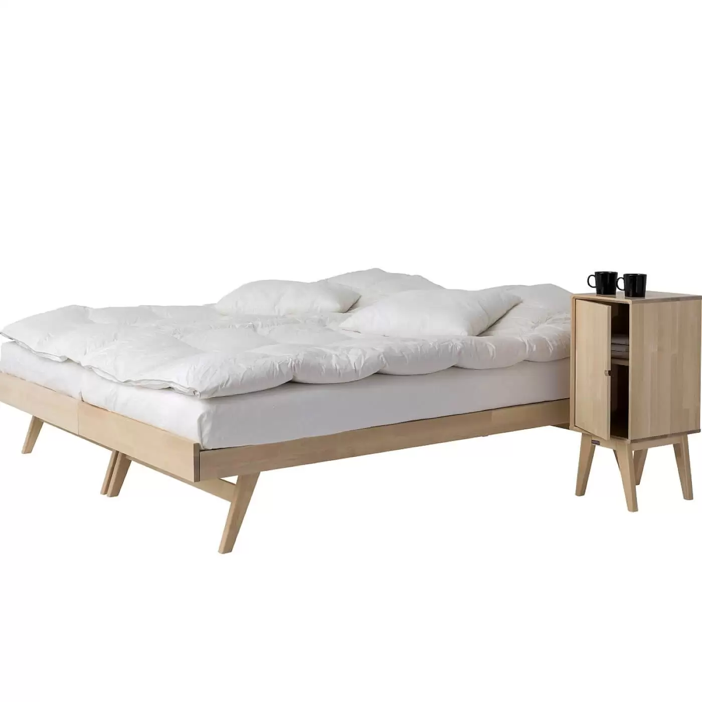 Łóżko na nóżkach NOTTE. Dwa łóżka drewniane z materacami i białą pościelą razem złączone. Obok stoi szafka nocna na nóżkach drewniana z uchylonymi drzwiczkami. Na szafce 2 czarne kubki