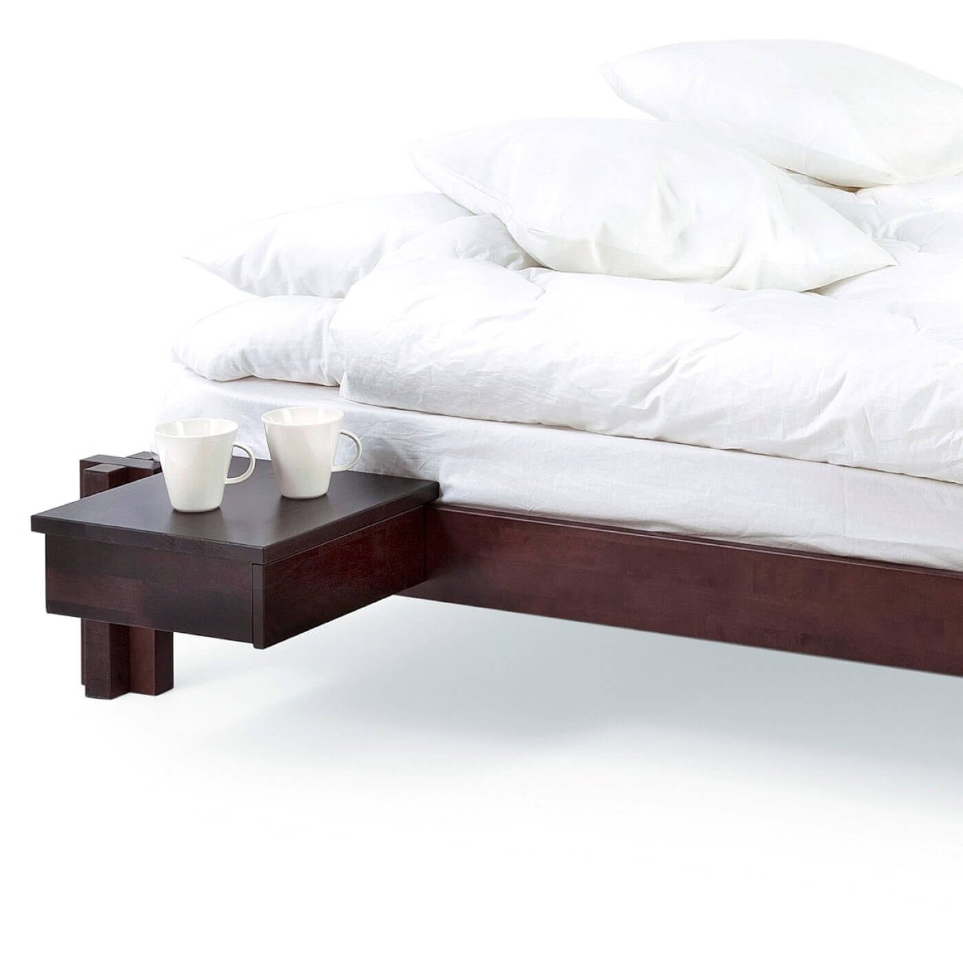 Niskie łóżko wenge MORI. Łóżko męskie nowoczesne widoczne we fragmencie z przywieszonym stolikiem nocnym w kolorze wenge z 2 białymi kubkami. Na łóżku z materacem leżąca biała pościel. Skandynawski design