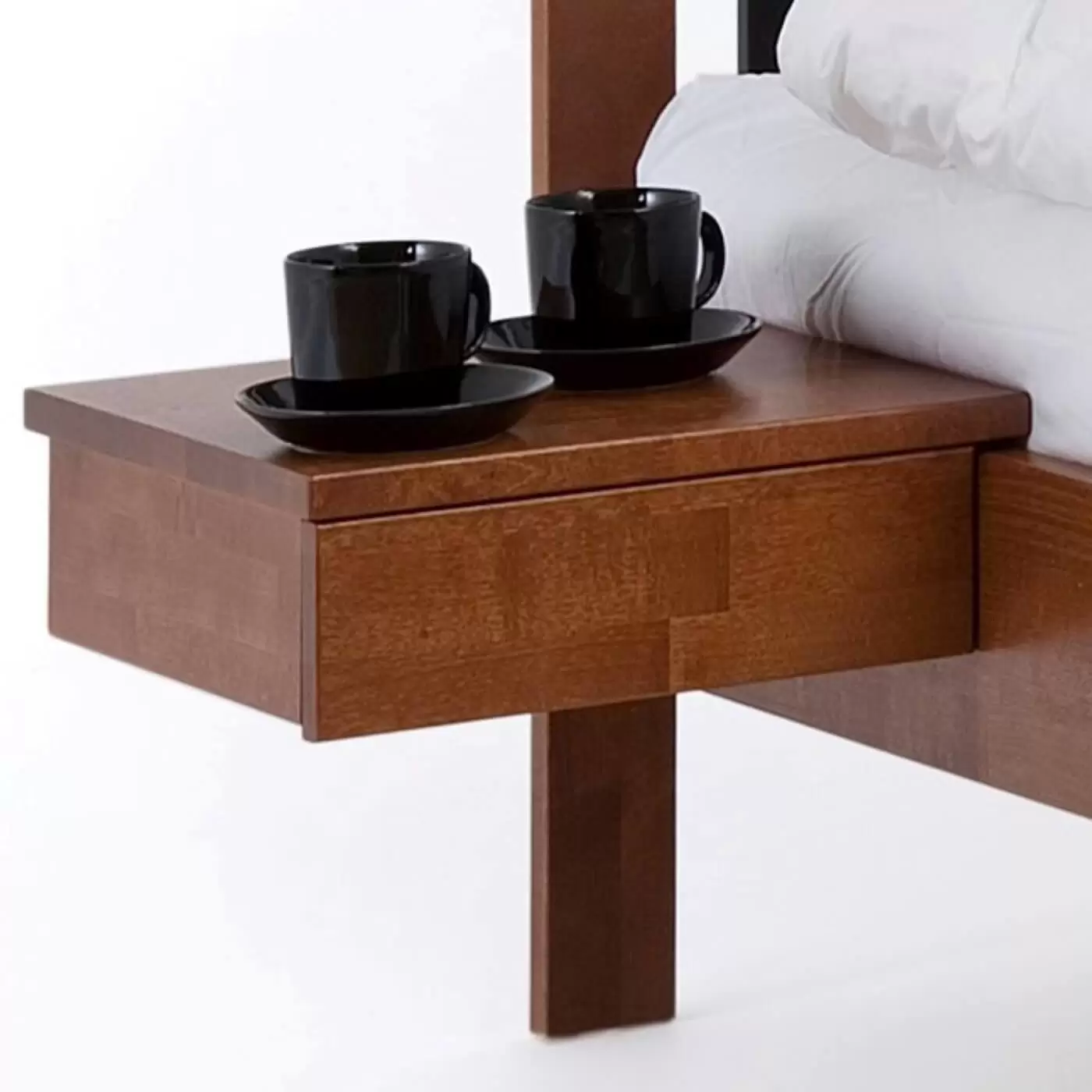 Stolik nocny orzech KOLI drewniany z szufladą. Przywieszony przy łóżku widoczny w dużym przybliżeniu. Na stoliku wiszącym z drewna w kolorze orzech widać stojące 2 czarne filiżanki. Nowoczesny skandynawski design