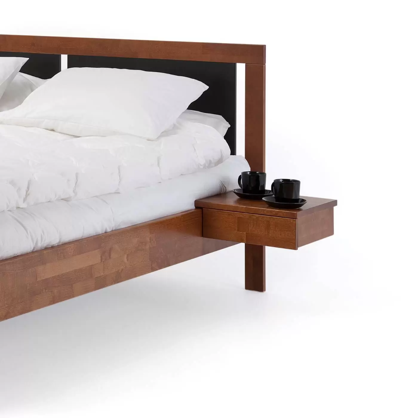Stolik nocny orzech KOLI drewniany z szufladą. Przywieszony przy łóżku na którym leży biała pościel. Na stoliku wiszącym orzech stoją 2 czarne filiżanki. Skandynawski design