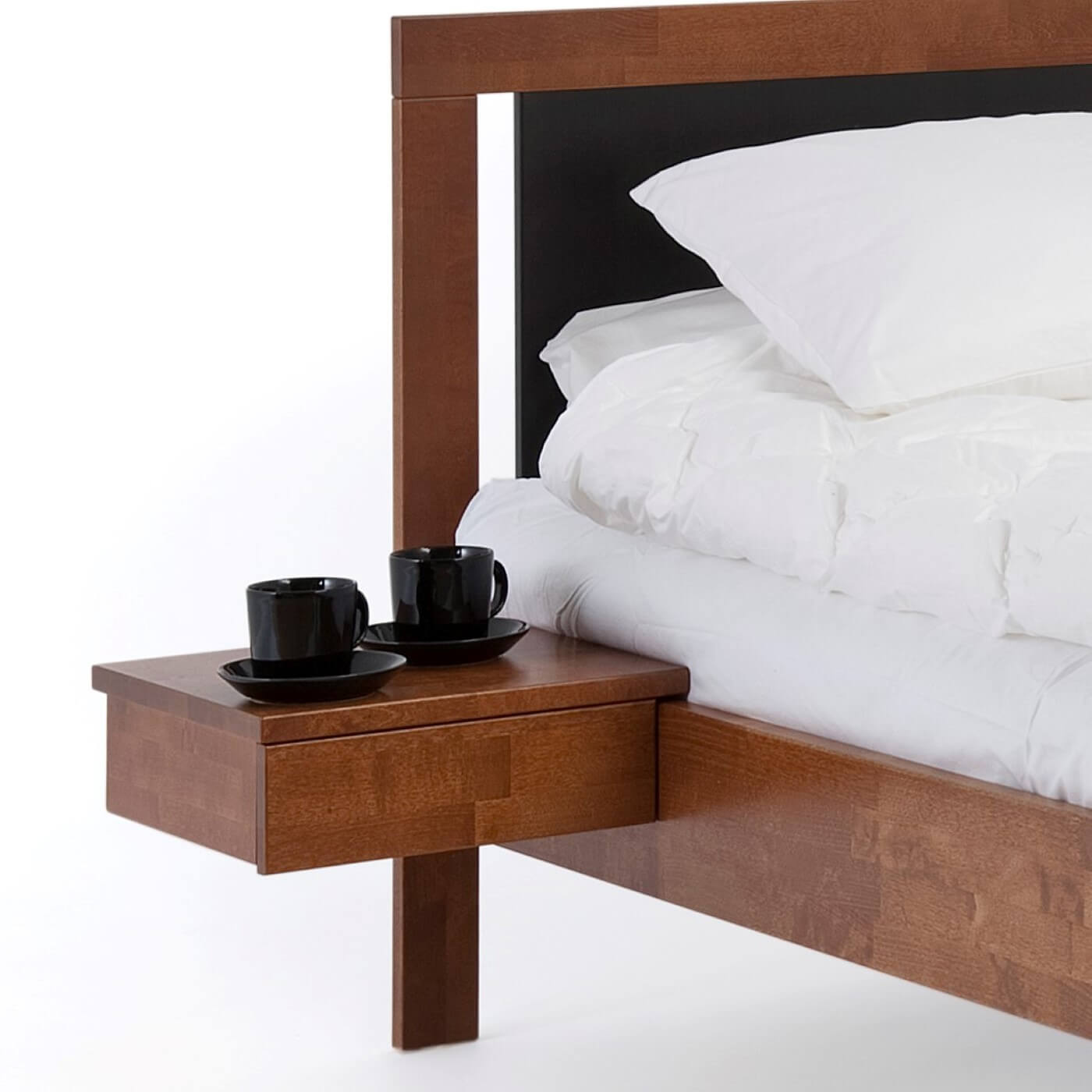 Stolik nocny orzech KOLI drewniany z jedną szufladą. Na stoliku wiszącym przy łóżku stoją 2 czarne filiżanki. Design z północy