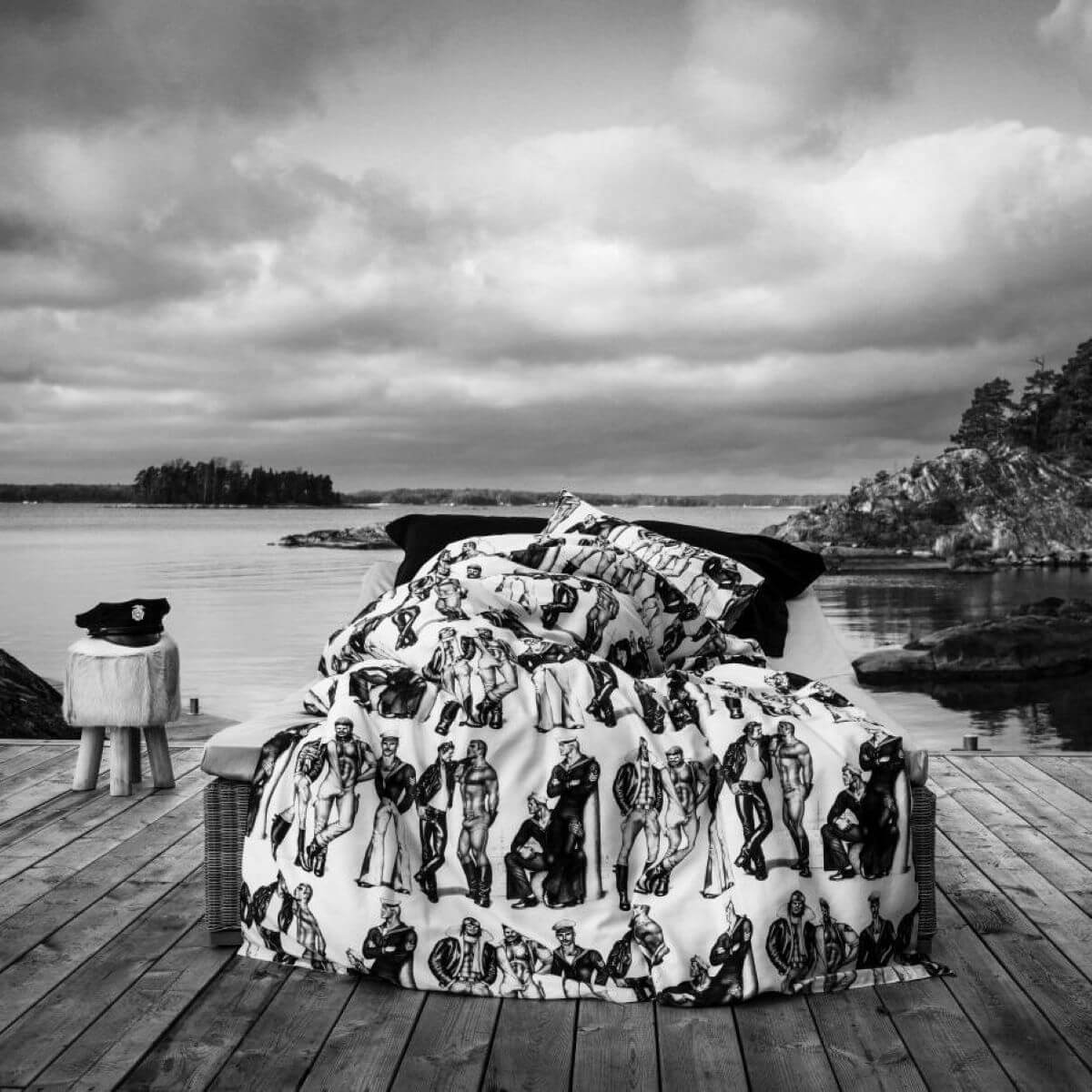 Pościel satynowa TOM OF FINLAND w czarno białej aranżacji na pomoście nad skandynawskim jeziorem. Biało czarna pościel satyna bawełniana rozłożona na łóżku. Wzór w pięknie zbudowane i zadbane męskie sylwetki w kolorach szaro czarnych na białym tle. Skandynawski design