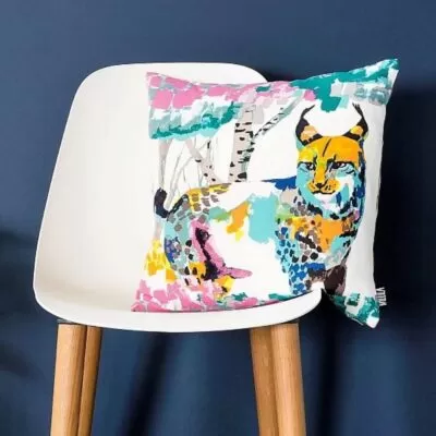 Poszewka dekoracyjna kolorowa ILVES. Bawełniana poszewka na poduszkę z kolorowym rysiem leżąca na nowoczesnym biało drewnianym krześle skandynawskim