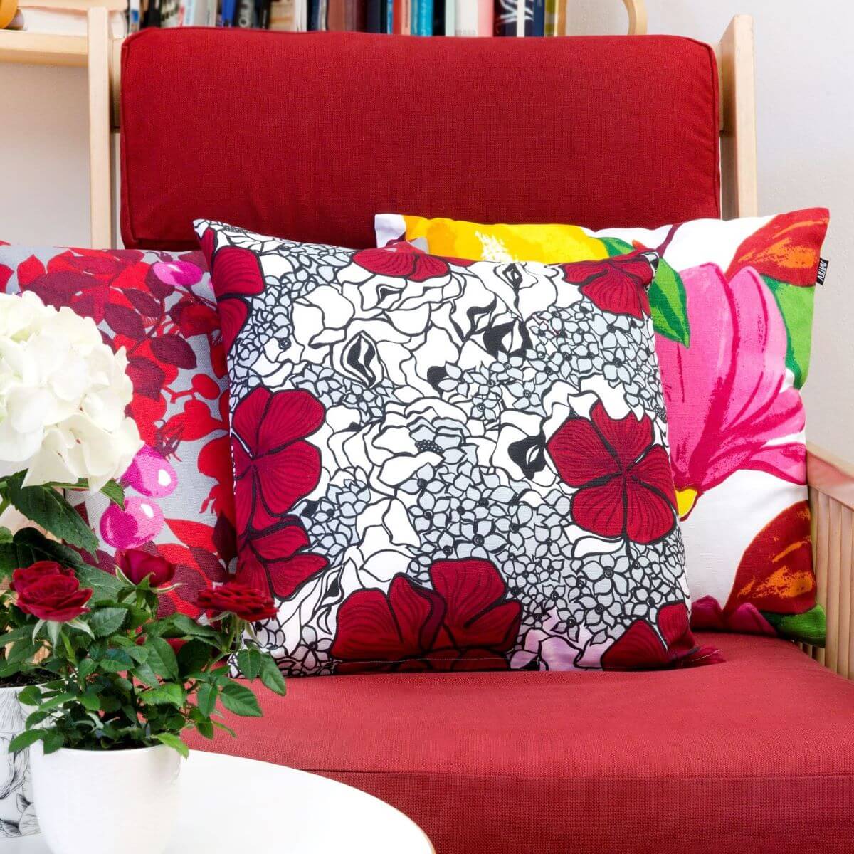 Poduszka dekoracyjna czerwone kwiaty ELLE. Leżąca na czerwonym fotelu poduszka z bawełny w czerwone i drobne szare kwiaty. Obok niej inne poduszki dekoracyjne w kwiaty. Skandynawski design