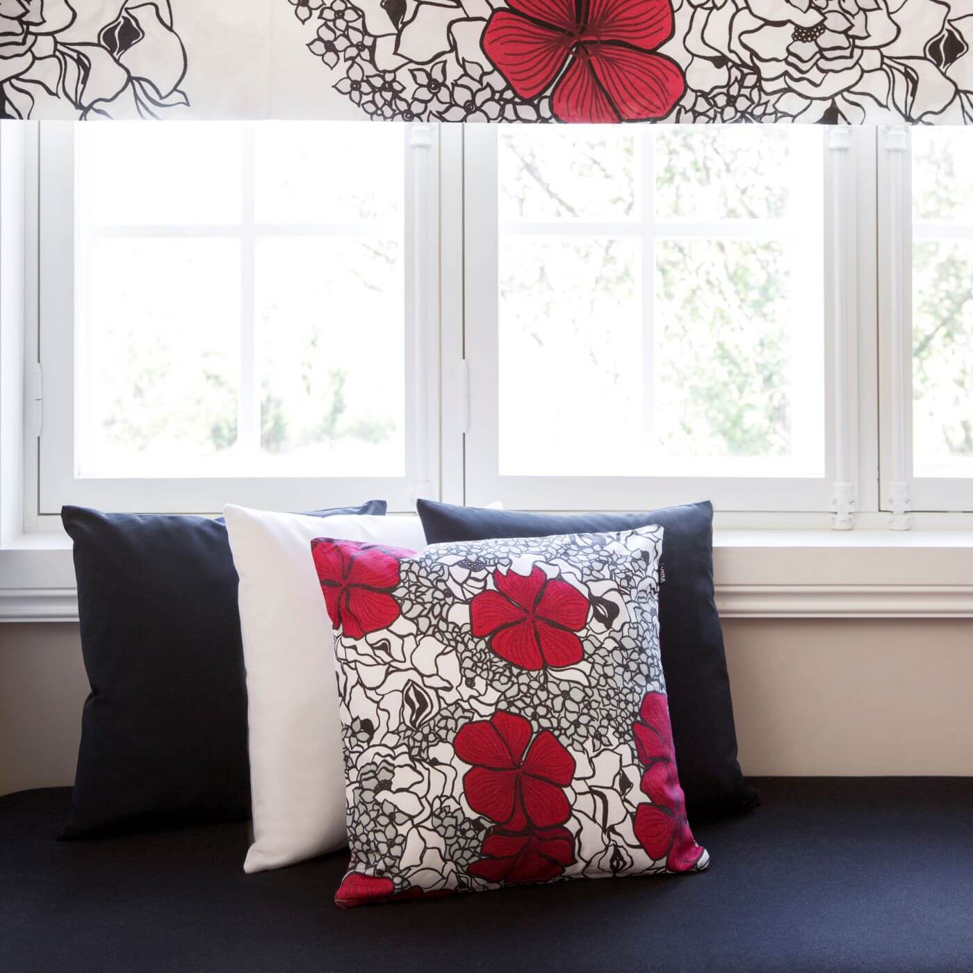 Poduszka dekoracyjna czerwone kwiaty ELLE. Bawełniana poduszka ze wzorem w splecione eleganckie czerwone i drobne szare kwiaty oraz 3 inne poduszki dekoracyjne leżące przy białym oknie. Skandynawski nowoczesny design