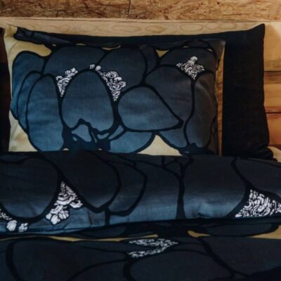 Poszewka elegancka MAKEBA. Widoczna w zbliżeniu bawełniana poszewka na poduszkę w duże kwiaty w szarym odcieniu leżąca na łóżku. Skandynawski design