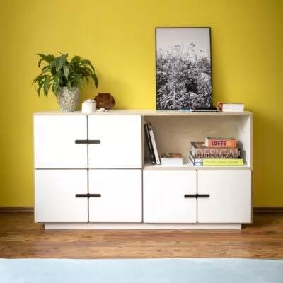 Komoda młodzieżowa PIX 2×4 ze sklejki pokrytej białym laminatem z półką i 6 drzwiami bez wystających uchwytów. Na tle żółtej ściany komoda z książkami obrazem i kwiatem. Skandynawski design